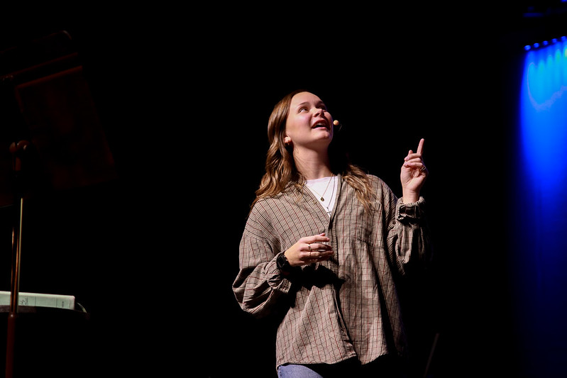 Female student speaking in chapel pointing up to the right to a screen not visible in the photo