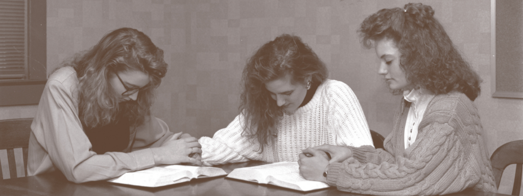 black and white photo of three female students praying in a classroom with bibles open in front of them.