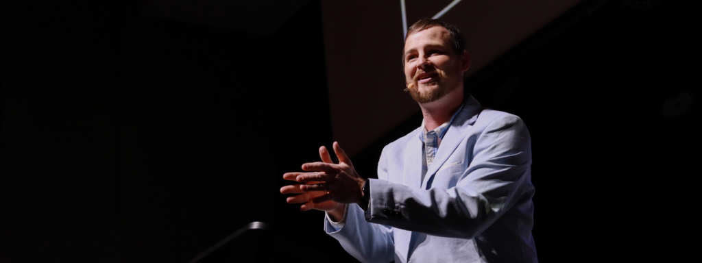 White male speaking in chapel with hands together pointing forward while smiling